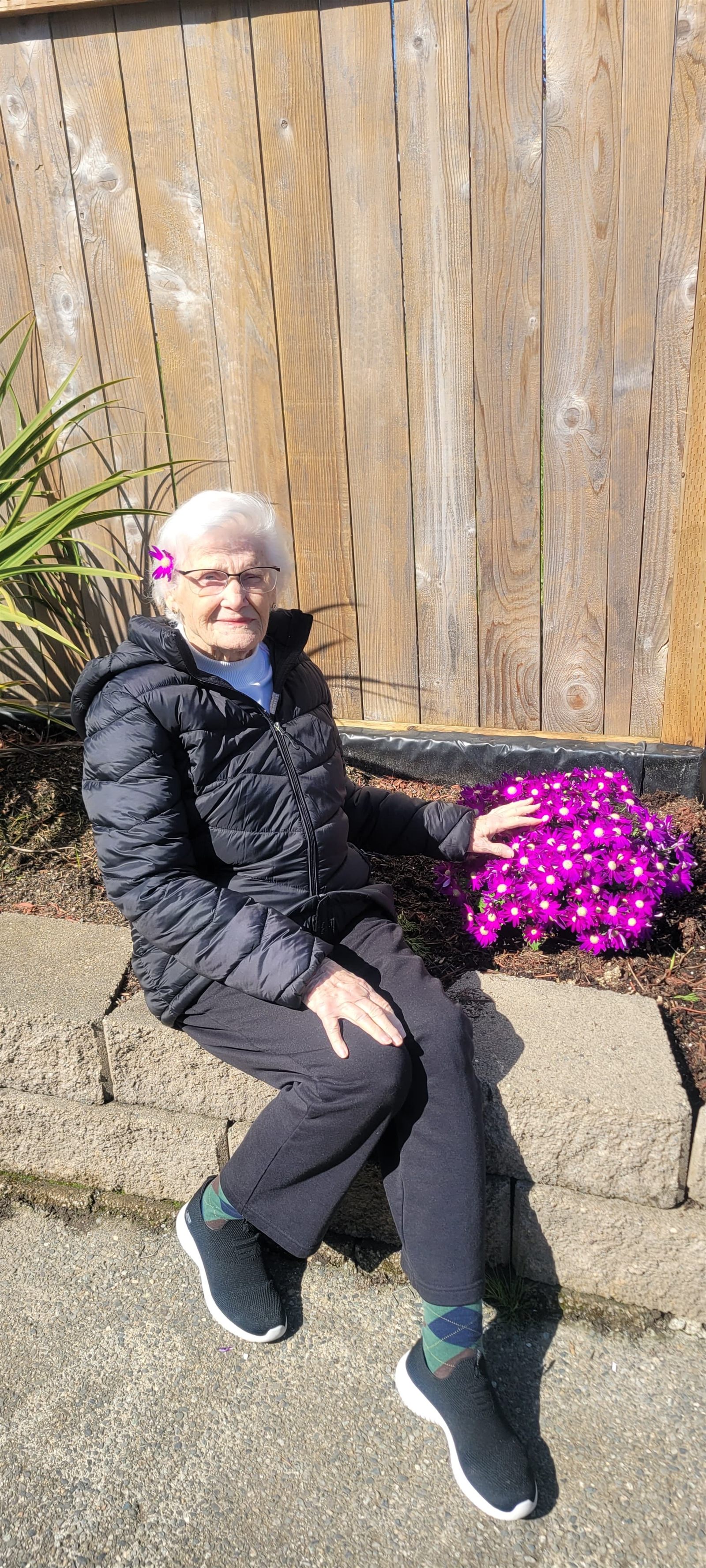 A resident sitting outside in the sunshine beside a bed of purple flowers